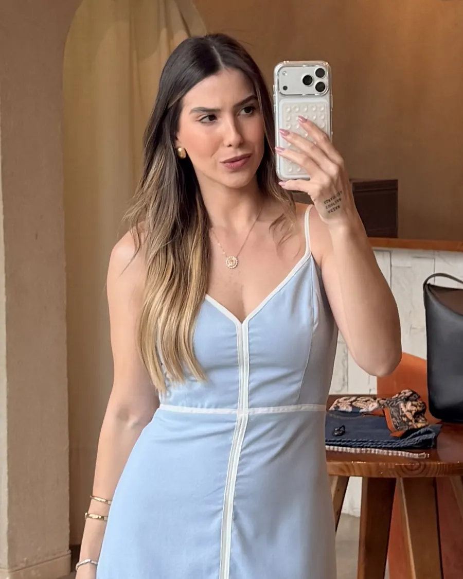 Ana Laura de vestido azul-claro, da cintura para cima, fotografando-se no espelho da loja