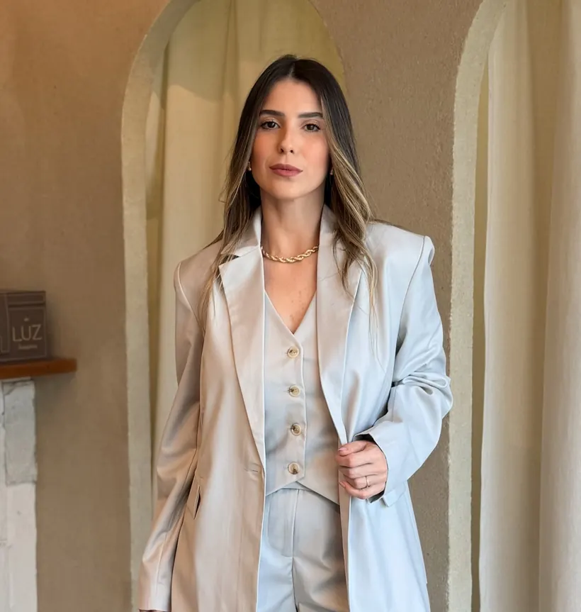 Ana Laura, dona da Lauss Concept, de conjunto de alfaiataria cinza-claro com blazer, de pé na loja, ao lado da cortina de linho